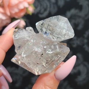 May include: A cluster of clear quartz crystals with a faceted, diamond-like shape. The crystals are clustered together in a natural formation, with some showing iridescent flashes of color.