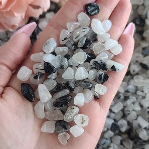 May include: A hand holding a pile of small, tumbled black and clear quartz stones. The stones are smooth and have a natural, polished look.