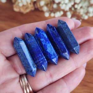May include: Five blue lapis lazuli crystal points, also known as wands, are arranged in a row on a hand. The points are smooth and polished, with a deep blue color and some white veining.