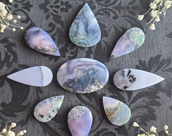 May include: Nine teardrop-shaped agate cabochons in shades of purple, grey, and green. The cabochons are arranged in a circular pattern on a dark grey background.