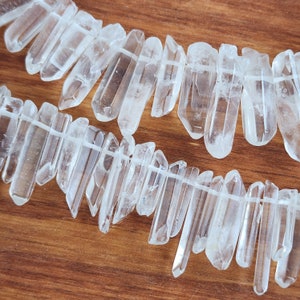 May include: Two strands of clear quartz crystal points, strung together with a white thread. The crystals are elongated and vary in size, with natural imperfections. The background is a warm, wooden surface.