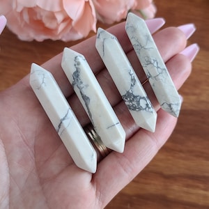 May include: Four white Howlite crystal wands with black veining. The wands are pointed at both ends and are arranged in a row on a hand.