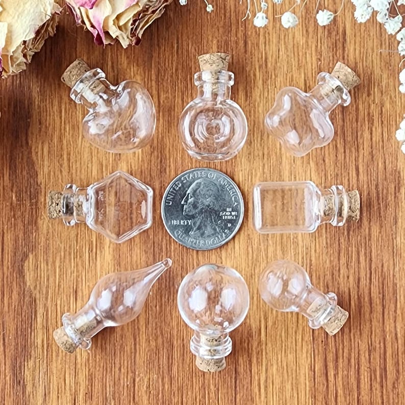May include: Assortment of nine small glass bottles with cork stoppers. The bottles are clear and come in various shapes, including round, heart, star, and rectangular. The bottles are arranged in a circle on a wooden surface.