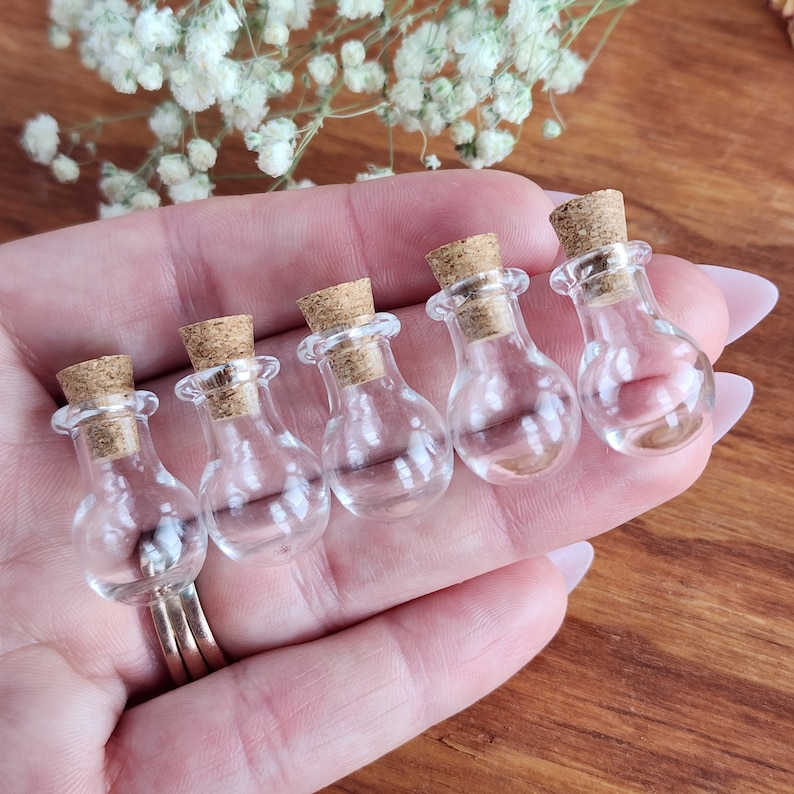 May include: Five small, clear glass bottles with cork stoppers. The bottles are round and have a slight bulb shape. They are arranged in a row, with the stoppers facing the viewer.