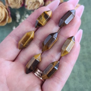 May include: Seven golden tiger's eye points, each with a unique pattern of brown and gold, are displayed on a hand. The points are faceted and have a smooth, polished finish.