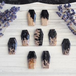 May include: A collection of ten coffin-shaped pendants with a black and tan marbled design. The pendants are arranged on a white wooden surface, with sprigs of lavender in the background. The pendants have a glossy finish.