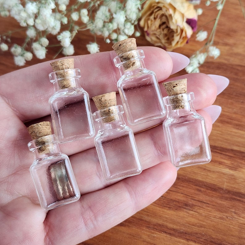 May include: Five small, clear glass bottles with cork stoppers. The bottles are square shaped and are arranged in a hand.