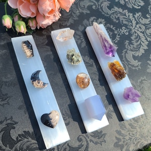 May include: Three white selenite charging plates with various crystals and stones placed on top. The crystals include amethyst, clear quartz, black moonstone, a brown and black heart, a green and brown cluster, a brown and black crescent moon, and a light blue crystal.