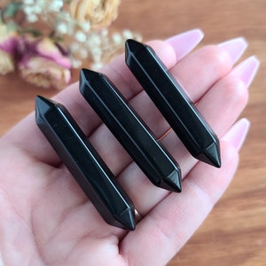 May include: Three black obsidian crystal wands, also known as pointing wands, are held in a hand. The wands are smooth and polished with a pointed tip.