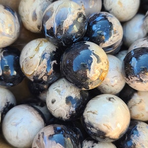 Può includere: Una collezione di sfere di pietra lucida nei toni del nero, bianco e beige. Le sfere hanno un aspetto marmorizzato con motivi a vortice e una finitura lucida. Le sfere sono disposte vicine, riempiendo l'inquadratura.