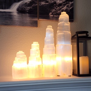 May include: Four selenite crystal lamps of varying sizes, arranged in a row on a shelf. The lamps are lit and emit a warm, white glow. The crystals are clear and have a natural, rough texture.
