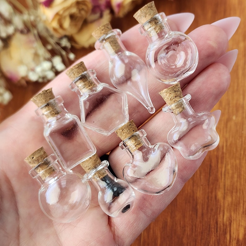 May include: A collection of nine small glass bottles with cork stoppers. The bottles are clear and have various shapes, including round, heart, and star.