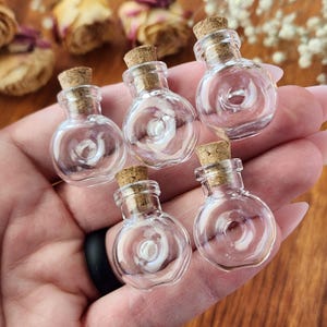May include: Six small, clear glass bottles with cork stoppers. The bottles are round and have a slight lip at the top. They are all empty and sitting on a hand.