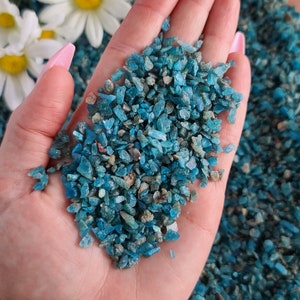 May include: A hand holding a pile of small, teal blue, green, and gray gemstone chips. The chips are irregularly shaped and have a sparkly, iridescent finish.