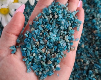 May include: A hand holding a pile of small, teal blue, green, and gray gemstone chips. The chips are irregularly shaped and have a sparkly, iridescent finish.
