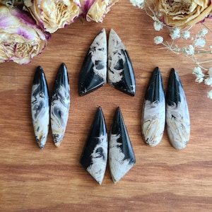 May include: A collection of polished gemstone cabochons in various shapes, including teardrop and triangular forms. The stones feature a striking contrast of black and cream-colored swirling patterns. The cabochons are arranged on a wooden surface.