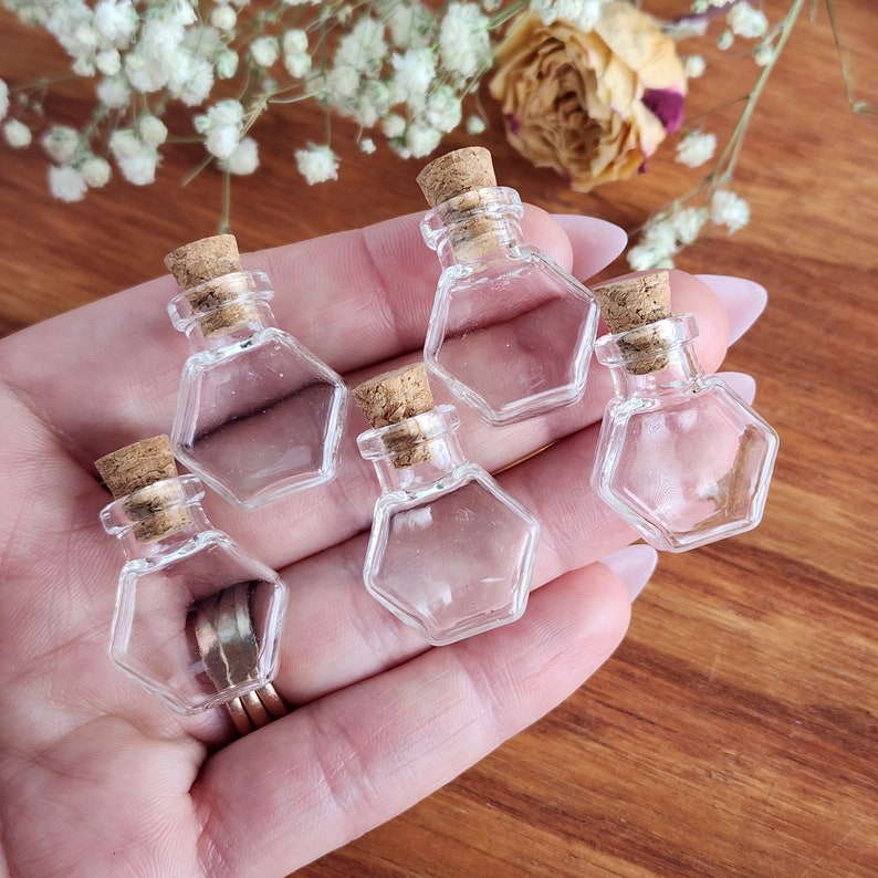 May include: Five small, clear glass bottles with cork stoppers. The bottles are hexagonal in shape and are held in a hand.