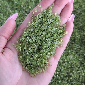 May include: A hand holding a pile of small, green, translucent gemstone chips. The chips are irregularly shaped and have a slightly rough texture.