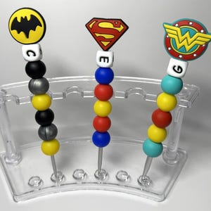 May include: Three colorful beaded necklaces with superhero character toppers. The necklaces are displayed on a clear plastic stand. The toppers are Batman, Superman, and Wonder Woman. The beads are yellow, red, blue, turquoise, black, and silver.