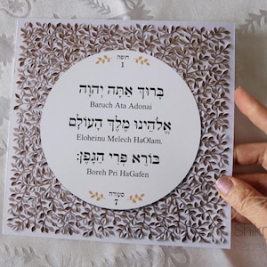 May include: A square, white paper card with a laser-cut leaf design in brown. The center features a white circle with Hebrew text and English transliteration. The card is held by a hand.