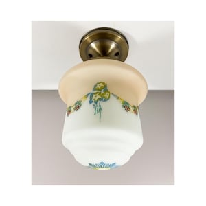 Può includere: Una lampada da soffitto con un paralume in vetro smerigliato a forma di campana, con una sfumatura dal pesca al bianco. Il paralume è decorato con una ghirlanda floreale dipinta in blu, giallo e rosso, fissata a una base circolare in ottone antico.