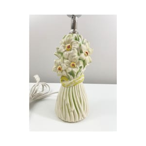 May include: A ceramic table lamp featuring a bouquet of white flowers with yellow centers and green leaves. The flowers are tied with a yellow ribbon. The base is a light green color with vertical lines. The lamp has a silver-colored metal neck and a white electrical cord.