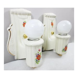 Könnte beinhalten: Zwei Vintage-Wandleuchten aus weißer Keramik mit Blumendesigns und goldenen Akzenten. Jede Wandleuchte hat eine runde, weiße Glühbirne. Die Wandleuchten haben ein dekoratives, verziertes Design mit einem Blumenmuster in Rot, Lila und Blau.