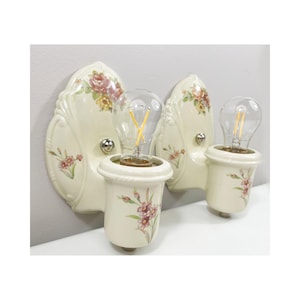 Könnte beinhalten: Zwei cremefarbene Vintage-Wandleuchten. Jede Leuchte hat ein Blumendesign mit rosa und roten Rosen und einer klaren Glühbirne. Die Leuchten sind aus Keramik und haben einen klassischen, eleganten Stil. Ca. 20 cm hoch.