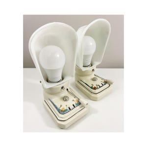 Może przedstawiać: Para kremowych lamp stołowych z ceramiczną podstawą, białym zakrzywionym kloszem, centralną białą żarówką oraz prostokątną podstawą ozdobioną kwiatowym wzorem w kolorach niebieskim, zielonym i pomarańczowym. Każda lampa posiada srebrny wyłącznik łańcuszkowy.