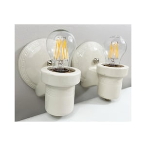 Könnte beinhalten: Zwei cremefarbene Keramik-Wandleuchten mit klaren Glühbirnen. Die Glühbirnen strahlen ein warmes gelbes Licht aus. Die Leuchten haben ein schlichtes, Vintage-Design, das sich perfekt eignet, um jedem Raum einen Hauch von Eleganz zu verleihen.