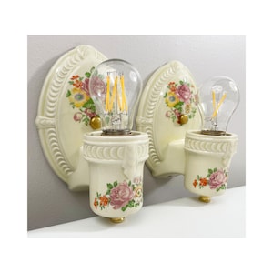 Könnte beinhalten: Zwei cremefarbene Wandleuchten aus Keramik mit floralen Mustern. Jede Leuchte hat eine dekorative Rückplatte, einen zylindrischen Sockel und eine klare Glühbirne. Das florale Design umfasst rosa, gelbe und orangefarbene Blumen.