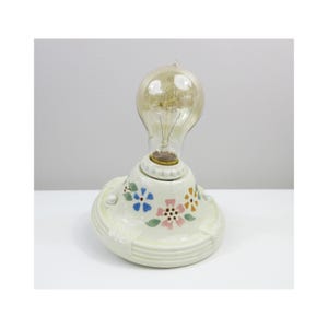 May include: A vintage-style lamp with a large, clear light bulb. The base is cream-colored ceramic, decorated with hand-painted flowers in blue, pink, and orange. The bulb is a classic Edison style.