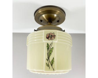 May include: A ceiling light fixture featuring a cylindrical cream-colored glass shade with a hand-painted pink flower and green stem design. The shade is attached to a circular, brushed brass-toned metal mount with a smooth, rounded finish.