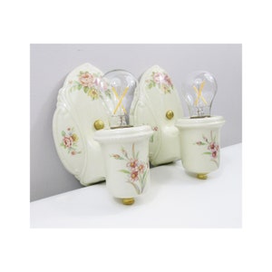 Könnte beinhalten: Ein Paar antike Wandleuchten aus Porzellan mit Blumendesigns. Jede Leuchte hat eine weiße Porzellanbasis mit einem Blumendesign und eine klare Glühbirne.