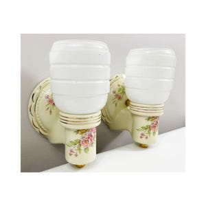 Könnte beinhalten: Paar Vintage-Wandleuchten. Jede Leuchte hat einen weißen, gerippten Glasschirm auf einem cremefarbenen Sockel. Der Sockel ist mit rosa und grünen Blumenmustern mit goldenen Akzenten verziert. Die Leuchten sind auf einer cremefarbenen Rückplatte montiert.