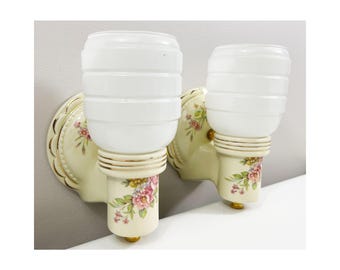 May include: Pair of vintage wall sconces. Each sconce features a white, ribbed glass shade atop a cream-colored base. The base is adorned with floral patterns in pink and green, with gold accents. The sconces are mounted on a cream-colored backplate.