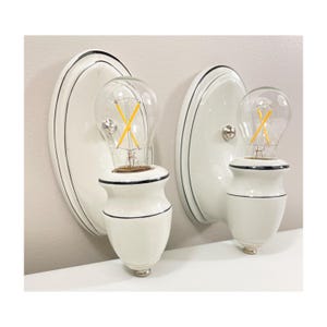 Könnte beinhalten: Zwei weiße Wandleuchten mit schwarzem Rand und klaren Edison-Glühbirnen. Die Leuchten haben ein klassisches Design mit einer abgerundeten Rückplatte und einem dekorativen Sockel. Die Glühbirnen haben einen warmen, gelben Schein.
