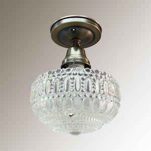 Pode incluir: Luminária de teto em estilo vintage com cúpula de vidro transparente estampada e componentes metálicos em latão antigo. A cúpula apresenta um design oval e pontilhado. A luminária é projetada para ser montada no teto.