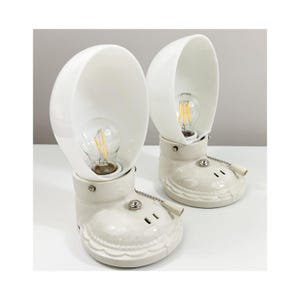 May include: Two ceramic lamps with a unique design. Each lamp features a white, semi-circular shade and a base shaped like a boot. The lamps have exposed light bulbs and pull chains for operation. The lamps are on a white surface.