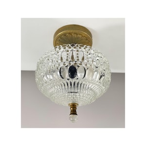Può includere: Una plafoniera a incasso con un paralume rotondo in vetro trasparente testurizzato con un motivo ripetitivo di ovali e punti. L'apparecchio include un rosone in metallo color ottone con un design a petali decorativo e un piccolo terminale in ottone.