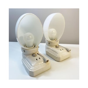 Könnte beinhalten: Zwei Vintage-Lampen aus cremefarbener Keramik mit einem abgerundeten, weißen Schirm und einer sichtbaren Glühbirne. Jede Lampe hat eine Zugkette und einen rechteckigen Sockel. Die Lampen sind für die Aufstellung auf einer ebenen Fläche konzipiert.