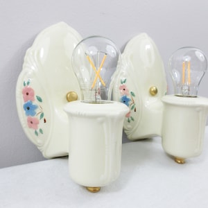 Könnte beinhalten: Cremefarbene Wandleuchten aus Keramik mit rosa und blauen Blumenmotiven. Jede Leuchte hat eine Glühbirne mit goldenem Sockel. Die Leuchten haben ein dekoratives, Vintage-Design, ideal, um jedem Raum einen Hauch von Eleganz zu verleihen.