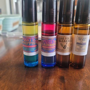 May include: Four essential oil roller bottles with black caps. The bottles contain colorful liquids and labels with text: "Dirty Hippie", "Fuzzy Rainbow", "Cure for the Bleakness", and "Away From The Mire". The bottles are on a wooden surface.