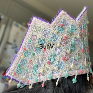 May include: A decorative crown with a diamond pattern in pastel colours, adorned with seashell and starfish charms. The crown has a purple trim and the text "SunV".