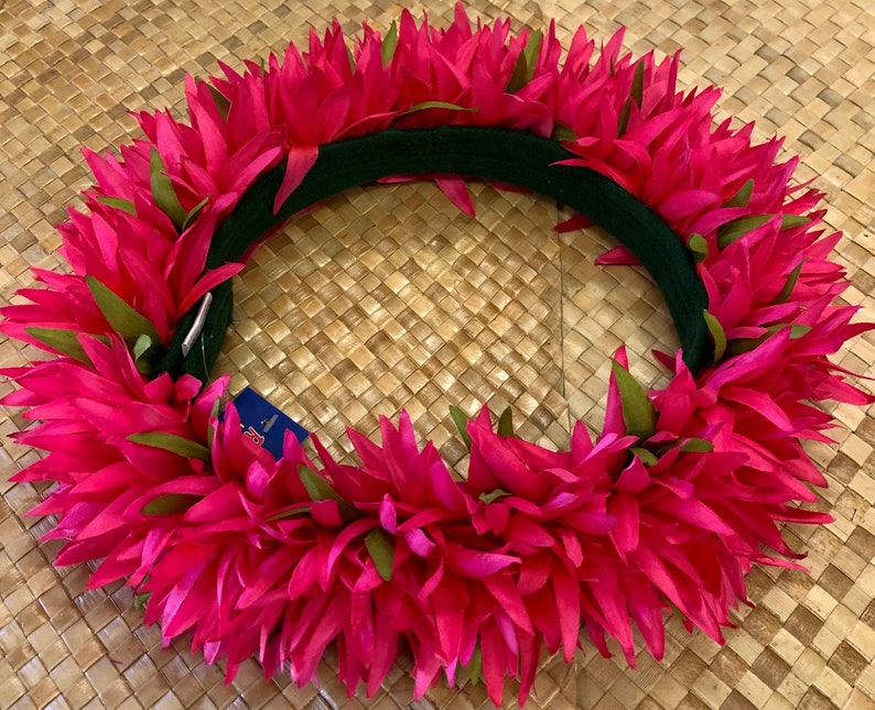Puede incluir: Un lei floral rosa vibrante con detalles verdes. El lei est&aacute; hecho de p&eacute;talos puntiagudos en capas y se coloca sobre una base verde oscuro. El fondo es una estera tejida de color marr&oacute;n claro.