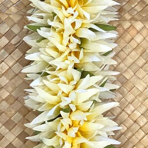 Puede incluir: Un lei vertical hecho de flores blancas y amarillas con acentos verdes. El lei se muestra sobre un fondo tejido de color marr&oacute;n claro. Las flores tienen un dise&ntilde;o de p&eacute;talos en capas.