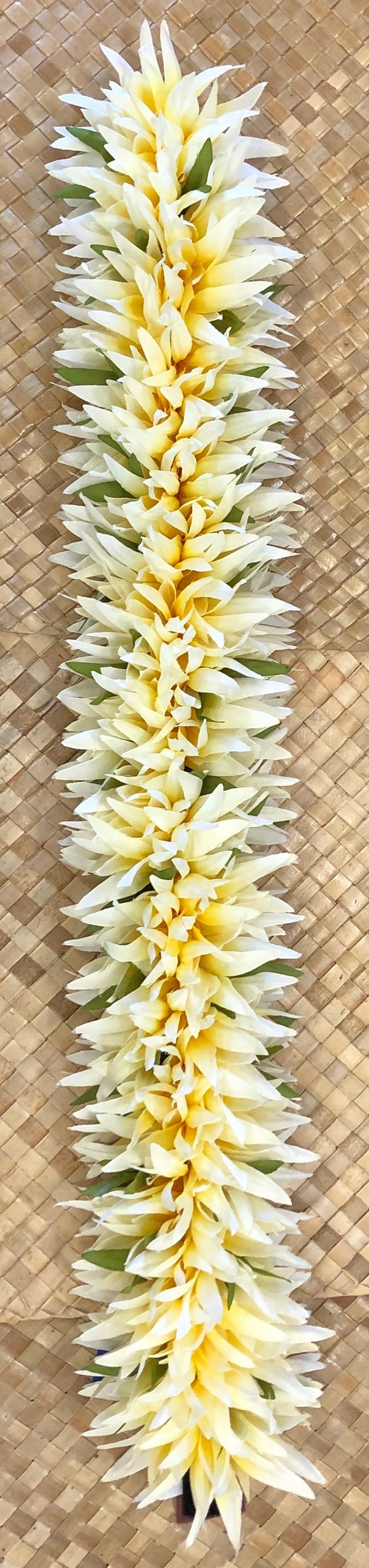 Puede incluir: Un lei vertical hecho de flores blancas y amarillas con acentos verdes. El lei se muestra sobre un fondo tejido de color marr&oacute;n claro. Las flores tienen un dise&ntilde;o de p&eacute;talos en capas.