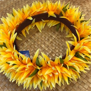 Puede incluir: Una vibrante corona floral amarilla y naranja con una base marr&oacute;n. La corona presenta capas de p&eacute;talos amarillos con acentos verdes, creando un dise&ntilde;o circular completo. La corona se encuentra sobre un fondo tejido.