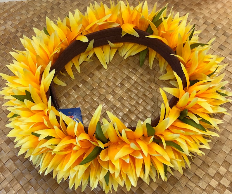 Puede incluir: Una vibrante corona floral amarilla y naranja con una base marr&oacute;n. La corona presenta capas de p&eacute;talos amarillos con acentos verdes, creando un dise&ntilde;o circular completo. La corona se encuentra sobre un fondo tejido.
