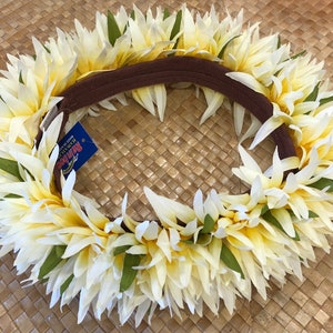 Puede incluir: Una vibrante corona de flores, con flores de color amarillo crema y blanco con detalles verdes. Las flores est&aacute;n dispuestas alrededor de una banda marr&oacute;n. Una etiqueta azul con texto est&aacute; adjunta. La corona se muestra sobre una superficie tejida.